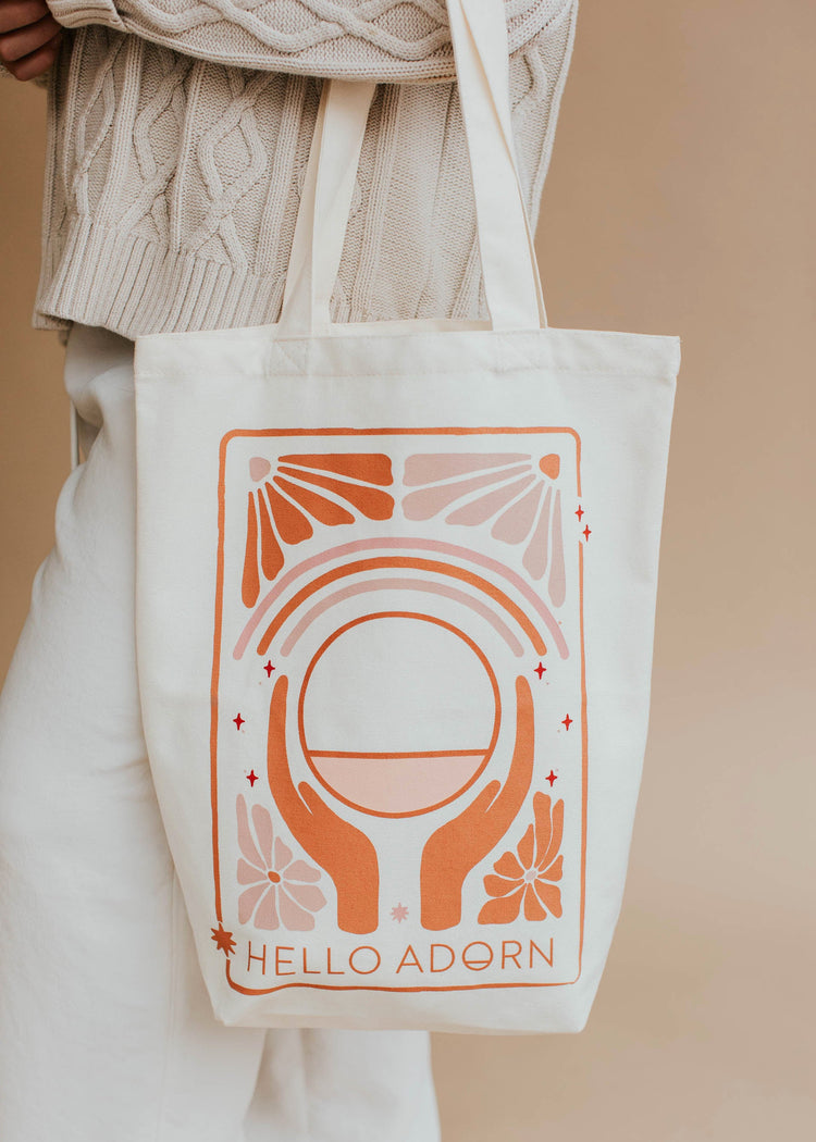 Tote bag with 'Hello Adorn' design held by a person wearing a beige sweater and pants.