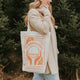 Woman holding a tote bag with a design and text, standing in a forest setting.