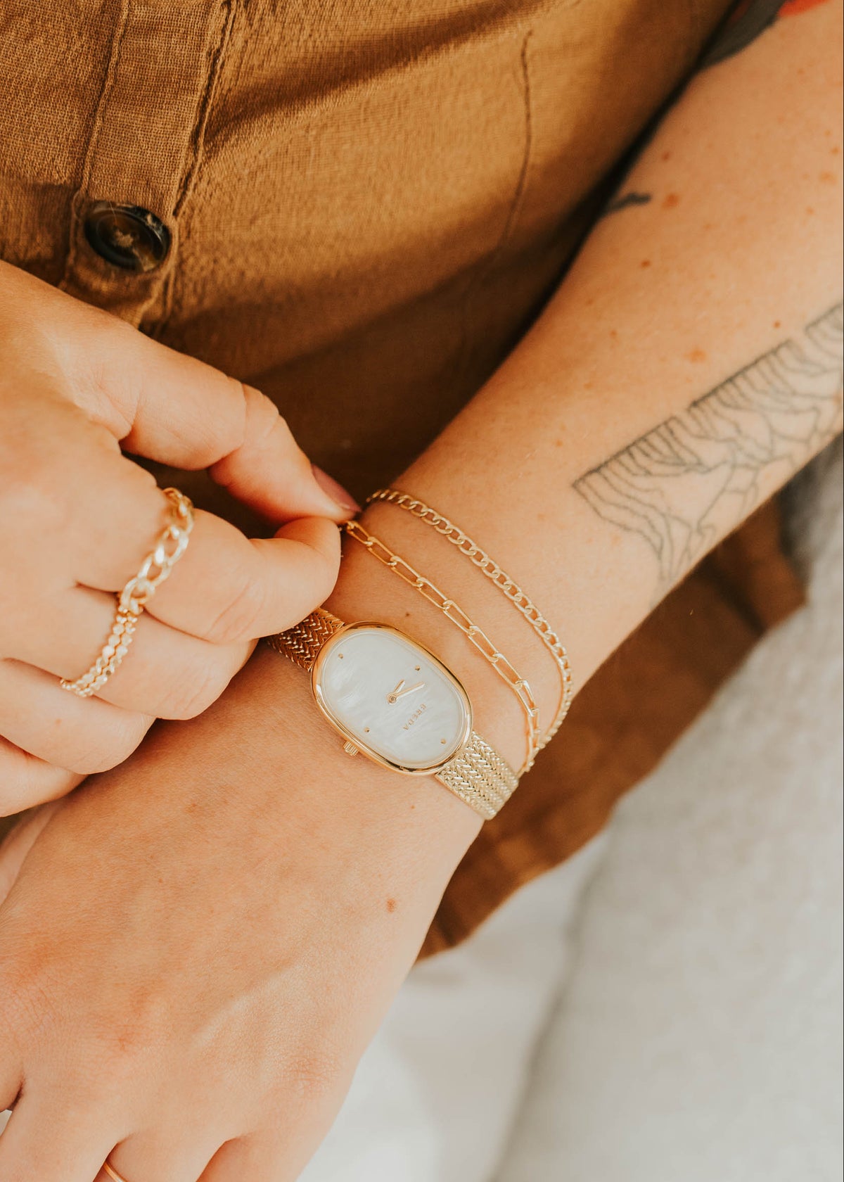 Close-up of hands wearing gold rings and a watch, with a brown garment in the background.