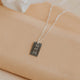 Silver necklace with a engraved tag on a beige surface with a white flower in the background