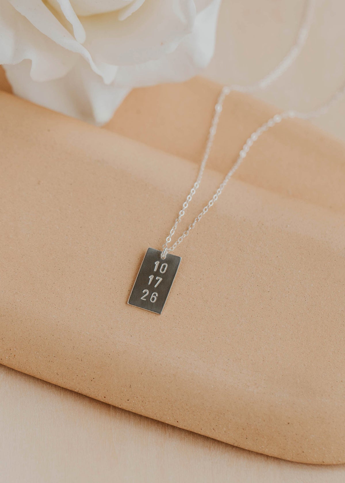 Silver necklace with a engraved tag on a beige surface with a white flower in the background
