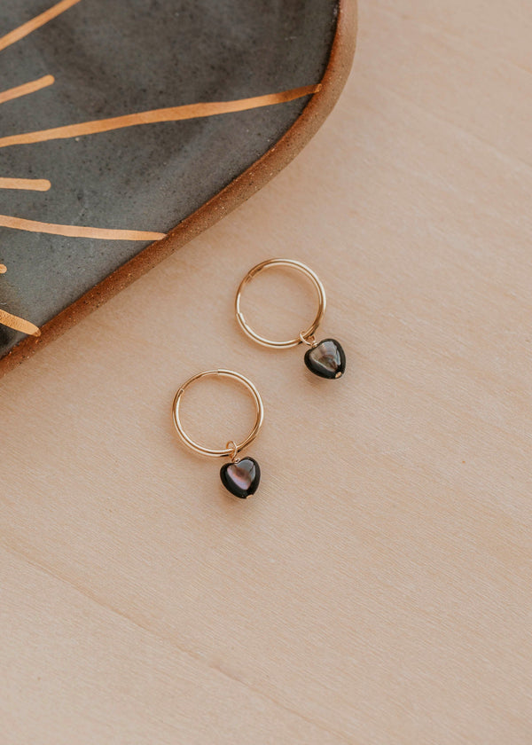 Gold hoop earrings with black heart charms on a wooden surface.