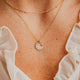 Close-up of a person wearing a Hello Adorn gold necklace with a pendant, with a white blouse in the background.