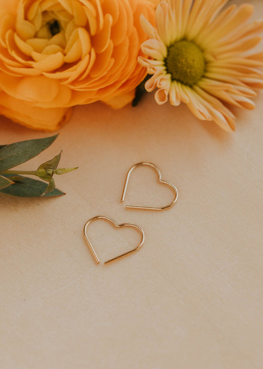 Gold heart-shaped earrings on a light surface with orange flowers and green leaves.